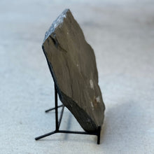 Load image into Gallery viewer, A dark gray slate stone display block on a small black metal stand, photographed on a light gray concrete surface.

